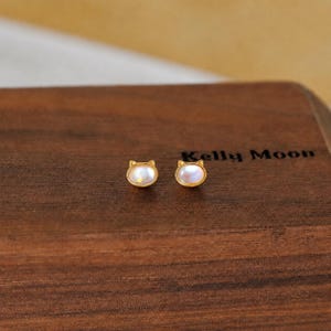 May include: A pair of gold-colored cat-shaped stud earrings with iridescent, oval-shaped stones. The earrings are displayed on a dark wooden surface, with the text "Kelly Moon" visible.