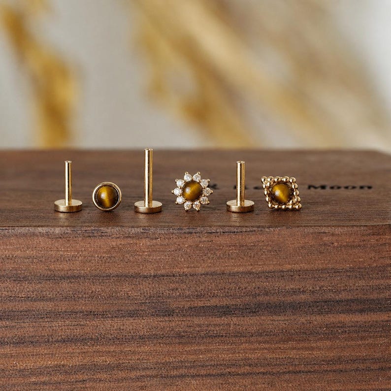 May include: A collection of gold-colored stud earrings with tiger's eye stones. The earrings feature various designs, including a round stone, a flower shape with surrounding crystals, and a square shape with a textured border. The earrings are displayed on a wooden surface.