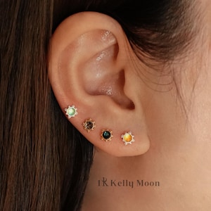 Op de afbeelding: Oor met vier oorstekers. De oorbellen hebben kleine, gekleurde edelstenen in groen, bruin, blauw en geel, elk omgeven door een decoratieve metalen zetting. De oorbellen zijn in het oor geplaatst, wat de sieraden laat zien.