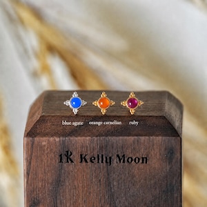 May include: Three gold-toned stud earrings featuring a central round gemstone, labeled blue agate, orange carnelian, and ruby. Each earring has a four-pointed star-shaped setting with small decorative beads. The text 1K Kelly Moon is engraved below.