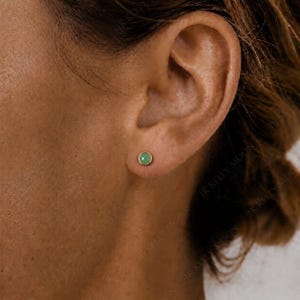 May include: A small, round stud earring featuring a smooth, light green cabochon stone set within a thin, polished gold-tone metal bezel. The earring is positioned centrally on the earlobe, showcasing a simple, circular design and a soft, matte finish.