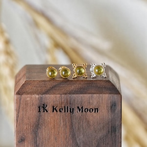 May include: Four pairs of stud earrings with olive-green gemstones. Two pairs are gold-toned, and two are silver-toned. The earrings are displayed on a wooden block with the text "1R Kelly Moon" engraved on the front.