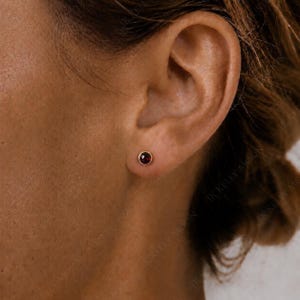 Può includere: Un piccolo orecchino a lobo rotondo con una pietra preziosa rosso scuro sfaccettata, incastonata in una montatura in metallo color oro lucido. L'orecchino è posizionato al centro del lobo, caratterizzato da un design essenziale e circolare.
