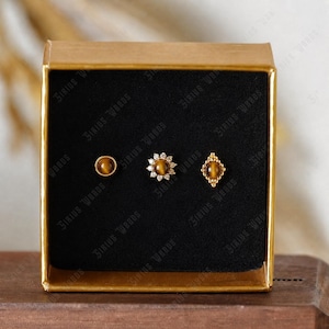 May include: A gold-toned box displaying three stud earrings. The earrings feature a round tiger's eye stone, a flower design with clear stones, and a diamond-shaped setting with a tiger's eye stone. The box has the text "Sirius Woods".