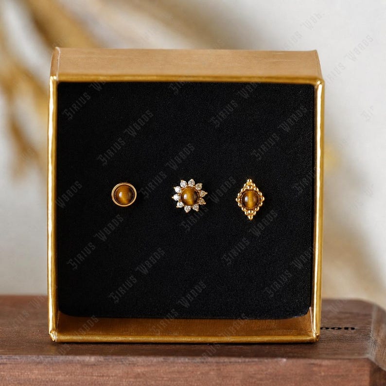 May include: A gold-toned box displaying three stud earrings. The earrings feature a round tiger's eye stone, a flower design with clear stones, and a diamond-shaped setting with a tiger's eye stone. The box has the text "Sirius Woods".