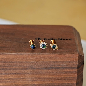 May include: Three gold-colored stud earrings with iridescent blue-green gemstones. The earrings are displayed on a dark wooden surface. One earring has a round stone, another has a starburst design, and the third has a square stone.