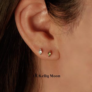 May include: A close-up of an ear with two small stud earrings. One earring has a light green stone, and the other has a white stone. The earrings are set in gold-colored metal. The text "1K Kelly Moon" is at the bottom.