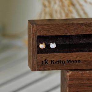 May include: A pair of cat-shaped stud earrings, one gold-toned and one silver-toned, displayed in a dark wooden jewelry box. The earrings are set against a dark velvet interior. The brand name "1R Kelly Moon" is visible on the box.