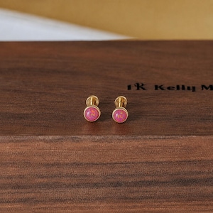 May include: A pair of gold-toned stud earrings featuring round, vibrant pink cabochon centers with subtle gold-colored flecks embedded within the surface. The earrings have a simple, polished metal post design with a circular bezel setting.