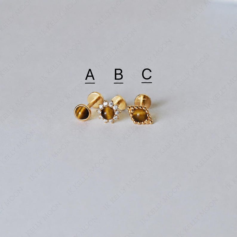 May include: Three gold-colored stud earrings with tiger's eye stones. The earrings are labeled A, B, and C. Earring A is a round stud. Earring B is a flower-shaped stud with small clear stones. Earring C is a diamond-shaped stud.