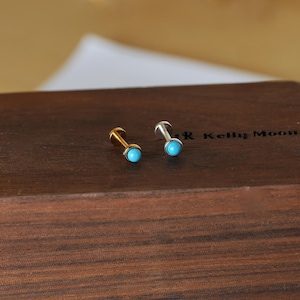 May include: Two small stud earrings with turquoise-colored stones. One earring has a gold-colored post and the other has a silver-colored post. The earrings are displayed on a dark brown wooden surface.