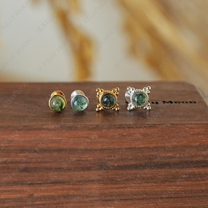 Puede incluir: Cuatro tapones para los oídos con centros de piedra verde. Dos son dorados, uno plateado y otro es un simple perno dorado. Los tapones se exhiben sobre una superficie de madera. El texto "IK KELLY MOON" es visible en el fondo.