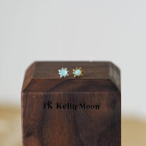 Op de afbeelding: Een paar oorknopjes, één zilver en één goud, elk met een lichtblauwe edelsteen in het midden en een zonnesterontwerp. De oorbellen liggen op een donkerhouten blok met de tekst "1R Kelly Moon" gegraveerd.