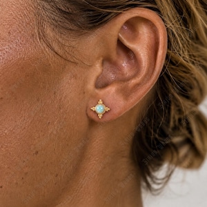 May include: A gold stud earring featuring a central round light blue opal stone surrounded by a decorative four-pointed star frame with beaded detailing. The earring is positioned in the earlobe, showcasing a polished metallic finish.