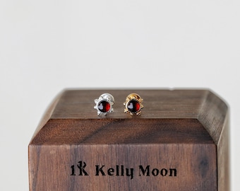 May include: A pair of stud earrings, one silver and one gold, each featuring a round, deep red gemstone. The earrings are displayed on a wooden block with the text "1R Kelly Moon" engraved on the front.