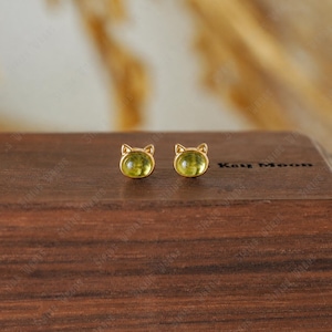 May include: A pair of gold-colored cat-shaped stud earrings. Each earring features a light green, rounded gemstone set within a gold frame. The earrings are displayed on a dark wooden surface, showcasing their design and color.