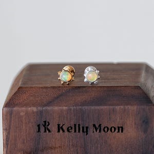 Könnte beinhalten: Zwei Ohrstecker mit schillernden Opalsteinen. Ein Ohrring ist goldfarben, der andere silberfarben. Die Ohrringe werden auf einem dunklen Holzblock mit dem eingravierten Text "1R Kelly Moon" präsentiert.