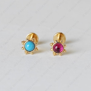 May include: Two gold-coloured stud earrings. One earring features a round, light blue stone, while the other has a round, pink stone. Both earrings have a decorative, sunburst-like design around the central stone.