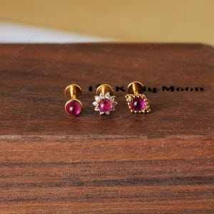 Könnte beinhalten: Drei goldfarbene Ohrstecker mit roten Edelsteinen. Die Ohrringe werden auf einer Holzoberfläche präsentiert. Ein Ohrring ist rund, einer hat eine Blütenform und der dritte ist rautenförmig.