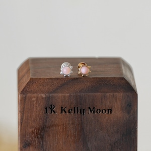 May include: Two stud earrings with pink gemstones, one silver and one gold, sit on a wooden block. The earrings have a floral design around the pink stone. The wooden block has the text "1R Kelly Moon" engraved on it.