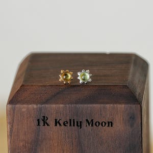 May include: A pair of stud earrings with a central gemstone, one gold-toned and one silver-toned, displayed on a dark wooden block. The earrings have a floral design around the gemstone. The wooden block is engraved with the text "1R Kelly Moon."