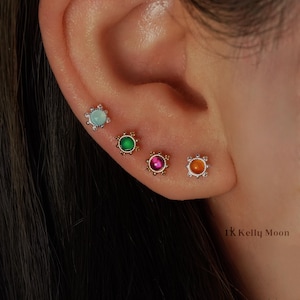 Op de afbeelding: Een oor met vier oorstekers. De oorbellen hebben gekleurde edelstenen in lichtblauw, groen, roze en oranje, elk omgeven door een decoratieve metalen zetting. De tekst "1K Kelly Moon" is zichtbaar. De oorbellen zijn klein.