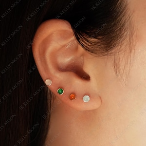 Könnte beinhalten: Nahaufnahme eines Ohrs mit vier Ohrsteckern. Die Ohrringe sind klein und haben farbige Edelsteine: weiß, grün, orange und weiß. Die Ohrringe sind in goldfarbenem Metall gefasst.
