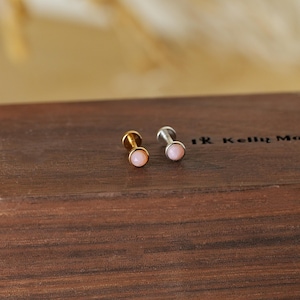 May include: Two small stud earrings with a gold and silver finish. Each earring features a round, light pink gemstone. The earrings are displayed on a dark brown wooden surface.