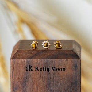 May include: Three gold stud earrings with tiger's eye stones are displayed on a wooden block. One pair is a simple round stud, another is a flower design with small white stones, and the third is a square design.