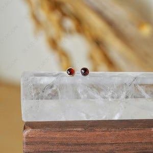May include: A pair of stud earrings featuring round, deep red gemstones set in gold and silver. The earrings are displayed on a clear, rectangular block, with a blurred, neutral background. The earrings are small and delicate.