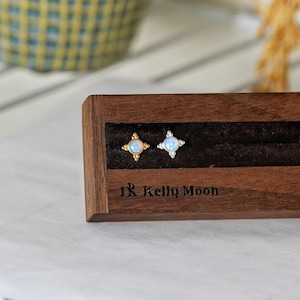 May include: Two stud earrings featuring round iridescent stones set in four-pointed star-shaped metal frames. One earring is gold-toned and the other is silver-toned. They are displayed in a dark brown wooden stand with the text Kelly Moon.