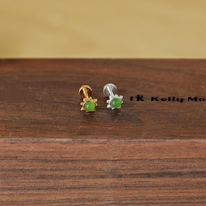 May include: Two stud earrings with a gold and silver finish. Each earring features a small, round, light green gemstone set in a decorative setting. The earrings are displayed on a dark wooden surface.