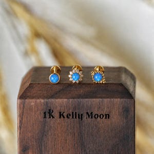 May include: Three gold stud earrings with round blue opal cabochons. The first is a simple bezel setting, the second has a sunburst frame with small clear stones, and the third has a rectangular beaded frame. The text 1K Kelly Moon is visible.