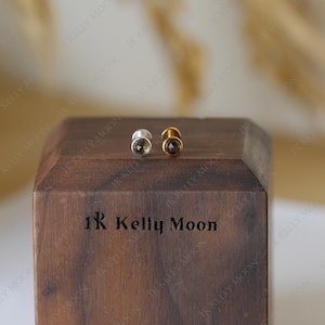 Puede incluir: Dos pendientes de botón con piedras de cuarzo ahumado. Un pendiente tiene un engaste plateado, el otro es dorado. Los pendientes se exhiben en un bloque de madera oscura con el texto "1K Kelly Moon".