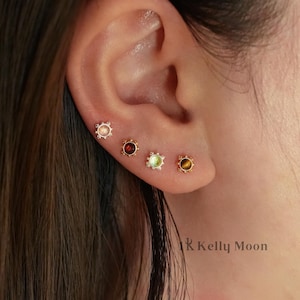 Pode incluir: Close-up de uma orelha com quatro brincos pequenos. Cada brinco apresenta uma pedra preciosa de cor diferente: rosa, vermelho, verde e castanho. Os brincos têm uma moldura dourada em forma de sol. A marca "Kelly Moon" é visível.