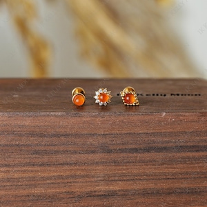 May include: Three gold stud earrings with orange gemstones. The earrings are displayed on a dark wooden surface. One earring has a round gemstone, another has a flower design with small clear stones, and the third has a square gemstone.