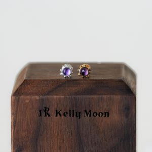 Puede incluir: Un par de pendientes de botón con una gema redonda de color púrpura en el centro. Un pendiente es plateado, el otro es dorado. Los pendientes se muestran en un bloque de madera oscura con el texto "1R Kelly Moon" grabado.