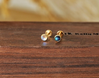 May include: Two gold-colored stud earrings. One earring features a round, iridescent, moonstone-like gem. The other earring has a round, dark blue, glittery gem. Both earrings are displayed on a dark brown wooden surface.