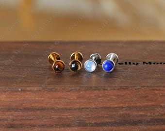 May include: A collection of stud earrings with various gemstones, including tiger's eye, black onyx, moonstone, and lapis lazuli. The earrings have gold and silver posts, displayed on a wooden surface. The gemstones are round and set in a bezel setting.