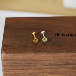 May include: Two stud earrings, one gold-toned with a yellow gemstone and one silver-toned with a green gemstone, rest on a dark wooden surface. The earrings have a simple, elegant design, perfect for everyday wear.