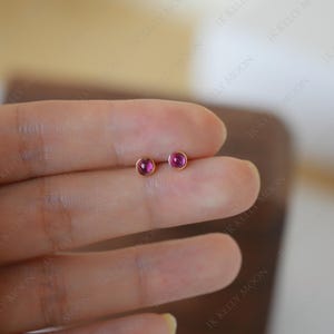 May include: A pair of small, round stud earrings. Each earring features a vibrant, pinkish-red gemstone set in a gold-colored bezel. The earrings are displayed on a light-toned background.
