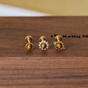 May include: Three gold-colored stud earrings with tiger's eye stones. One is round, one is a flower shape with small clear stones, and one is a diamond shape with a textured border. The earrings are displayed on a dark wooden surface.