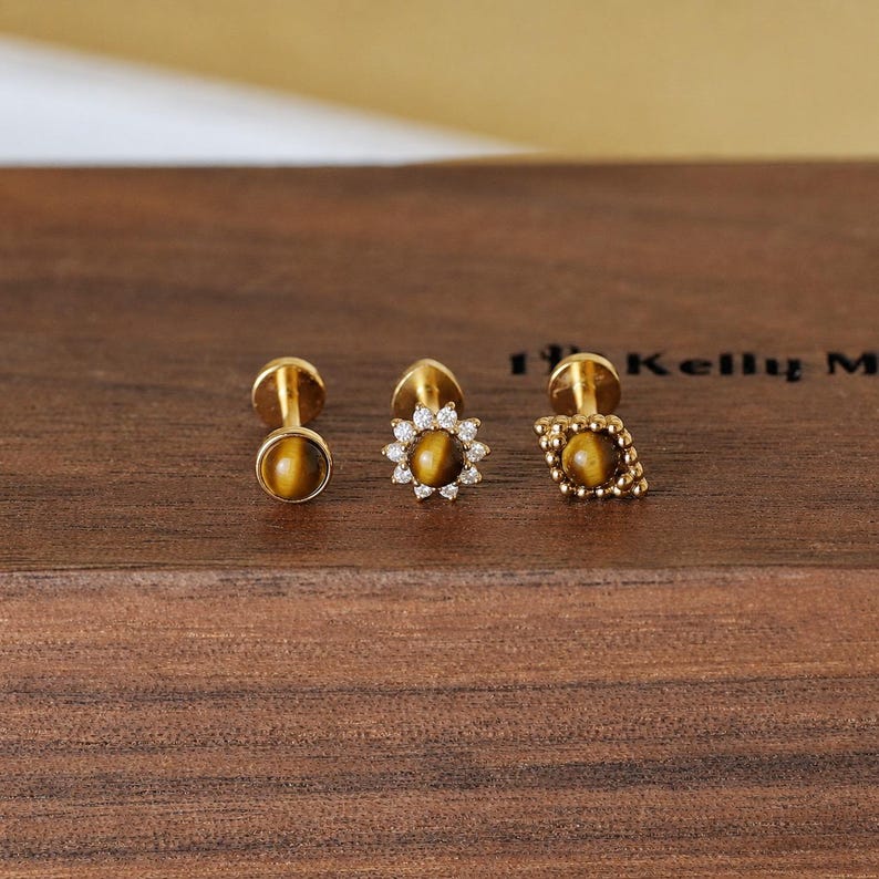 May include: Three gold-colored stud earrings with tiger's eye stones. One is round, one is a flower shape with small clear stones, and one is a diamond shape with a textured border. The earrings are displayed on a dark wooden surface.