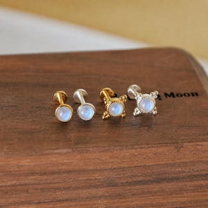 Puede incluir: Cuatro pendientes de botón con piedras redondas azul claro iridiscentes. Dos pendientes tienen postes dorados, uno tiene un poste plateado y otro tiene un engaste dorado decorativo. Los pendientes se exhiben sobre una superficie de madera.