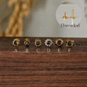 May include: Six gold-tone threaded stud earrings labeled A through F, featuring dark green stones. Design A has a beaded edge, B is a simple bezel, C is a textured bezel, D has a halo of clear stones, E is octagonal, and F has a decorative frame.