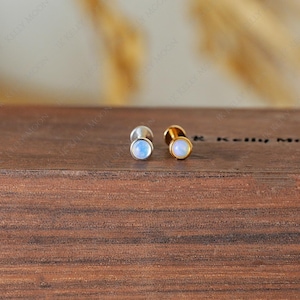 May include: Two stud earrings with round, iridescent moonstone cabochons. One earring has a silver setting, and the other has a gold setting. The earrings are displayed on a dark wooden surface.