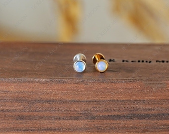 May include: Two stud earrings with round, iridescent moonstone cabochons. One earring has a silver setting, and the other has a gold setting. The earrings are displayed on a dark wooden surface.