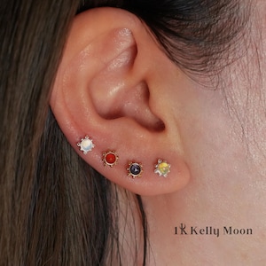 May include: Close-up of an ear adorned with four stud earrings. The earrings feature various gemstones in white, red, purple, and yellow, each set in a decorative, gold-toned setting. The text "1R Kelly Moon" is visible in the lower right corner.