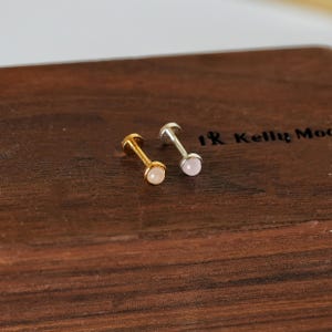 Op de afbeelding: Twee kleine oorstekers. Eén is goudkleurig met een lichtroze edelsteen, de andere is zilverkleurig met een lichtroze edelsteen. De oorbellen worden gepresenteerd op een donkerbruin houten oppervlak.