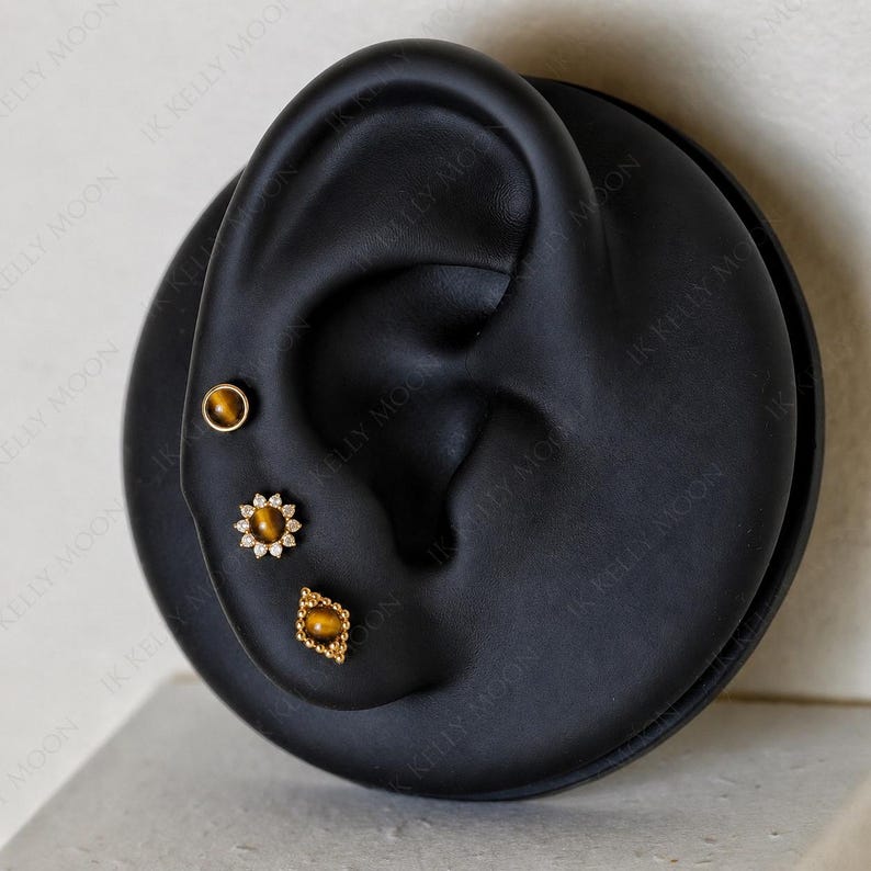 May include: Three gold-colored stud earrings with tiger's eye stones are displayed on a black ear-shaped display. One is a simple round stud, another is a flower shape with small clear stones, and the third is a diamond shape.
